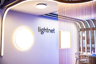 Illuminated Lightnet exhibition stand with round wall lamp, linear lighting, and seating area behind chain curtain.
