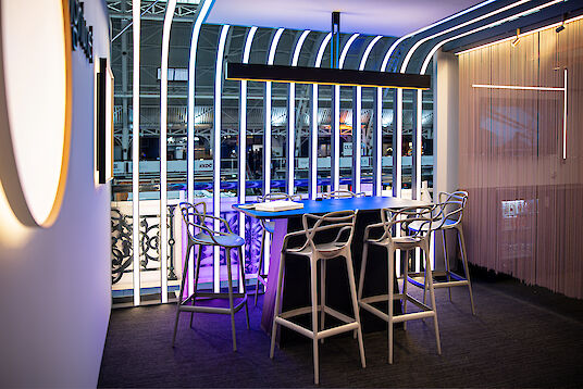 Modern illuminated exhibition stand with high chairs, table, and vertical light structures in the background.